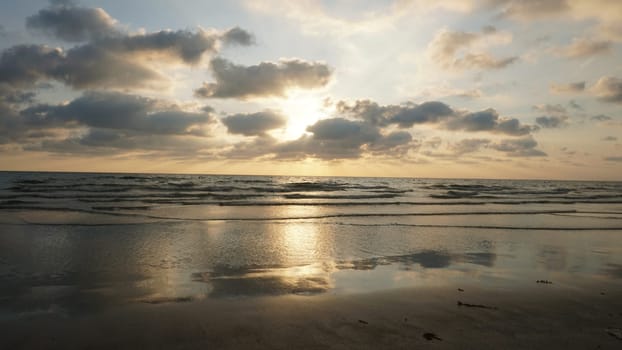 Soft sunlight filters through a cloudy sky, illuminating gentle waves rolling onto a wet sandy beach, creating a serene and reflective seascape at sunset
