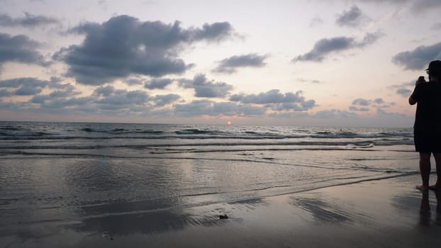 Photographer standing on a wet sandy beach at sunset, capturing vibrant sky colors reflected in gentle waves as the sun sets below the horizon