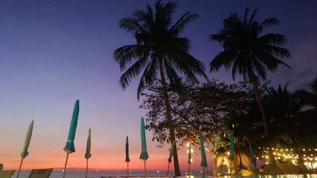 Closed beach umbrellas stand tall against a vibrant sunset sky, with silhouetted palm trees and string lights adding to the tranquil tropical atmosphere of a luxurious resort