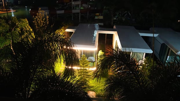 Warm lights illuminating modern bungalow at night, nestled among lush tropical foliage, with a water park shimmering in the background, creating a serene and inviting vacation atmosphere