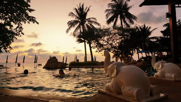 Tourists are enjoying a swim in a luxurious infinity pool adorned with elephant statues as the sun sets over the ocean, creating a tranquil and exotic atmosphere