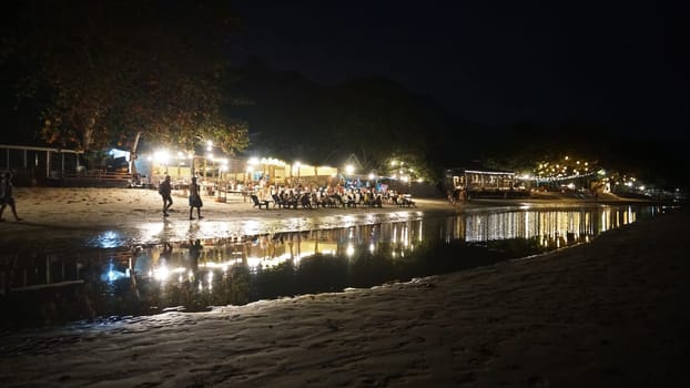 Lively beach restaurant at night with tourists dining and socializing, illuminated by warm lights reflecting in the tranquil water, creating a magical ambiance
