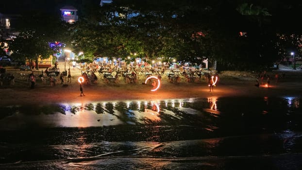 Street artists performing a fire show on a beach at night, with tourists dining at an outdoor restaurant under the trees and lights reflecting on the wet sand