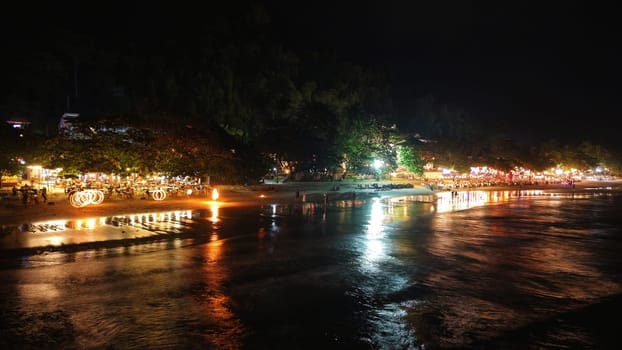 Tourists enjoying a vibrant fire dancing show on the beach, surrounded by restaurants at night, with colorful lights reflecting off the wet sand and shimmering sea
