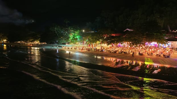 Tourists are dining and enjoying the vibrant atmosphere of a beachfront restaurant at night, with colorful lights reflecting on the wet sand and gentle waves lapping the shore