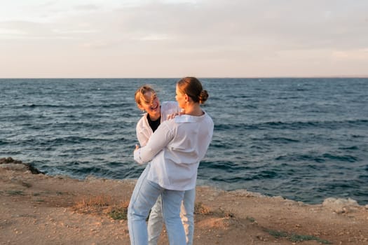 A couple stands on a cliff overlooking the ocean. The woman is holding the man in her arms. They are both smiling and looking at each other. The sun is setting in the background.