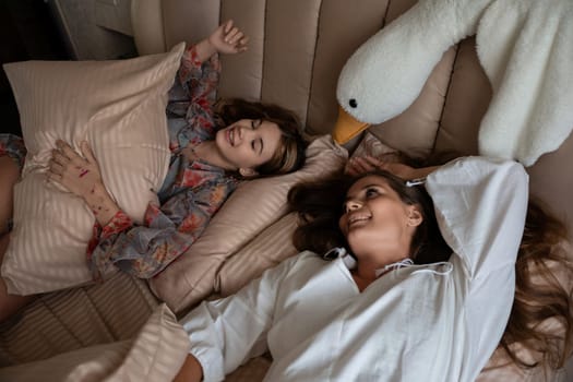 Two women lie in bed, smiling. One holds a pillow, the other rests, a large swan-shaped plush toy nearby.