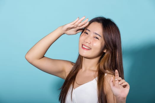 Asian young woman hand up cover face protect sun light and UV smile in studio shot isolated on blue background, female raising arm and hand in protection and block sunshine, sunscreen cream advertise