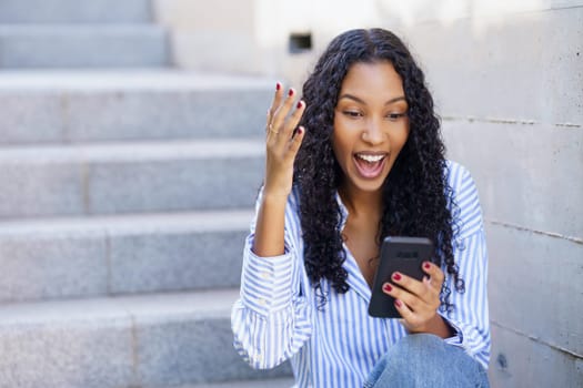 A joyful young woman who has beautiful curly hair is excitedly interacting with her smartphone while sitting on a set of steps, showcasing her happiness and connection to the digital world around her