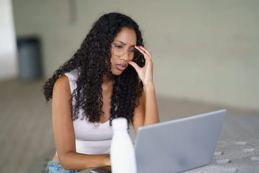 In a modern, contemporary environment, a woman with curly hair and stylish glasses is intensely concentrating on her laptop, showcasing her dedication and focus as she engages in task at hand