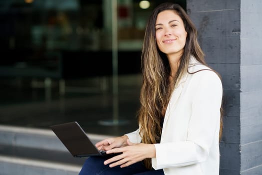 A young woman smiles confidently using her laptop outdoors in a vibrant urban setting, blending professionalism with casual style, illustrating the intersection of technology and creativity