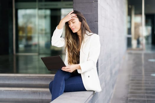 A young woman, elegantly dressed in a trendy outfit, sits comfortably on office steps, intently using her laptop, showing dedication and determination in her work and career pursuits