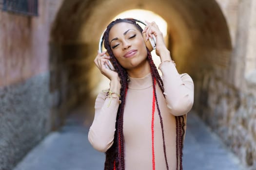 A young woman with braided hair enjoys music through stylish headphones in a narrow alleyway, radiating joy and confidence, embodying a carefree lifestyle that inspires and reflects urban culture