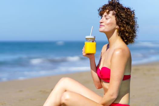 Calm curly female in bikini having glass cup of juice with straw while enjoying sunlight on sandy beach of ocean