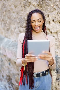 A cheerful woman possessing long, beautifully braided hair is deeply engaged with her tablet, relishing her leisure time in the picturesque outdoors, leaning against a beautifully textured wall