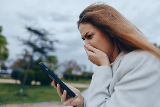 An anxious woman in casual clothes reacts with worry and stress to her phone content outdoors, cloudy sky behind, symbolizing financial crisis and emotional tension.