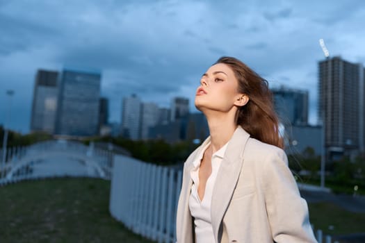 confident woman wearing beige coat and white top poses thoughtfully outdoors during dusk with city buildings behind, autumn fashion style with cool blue evening sky