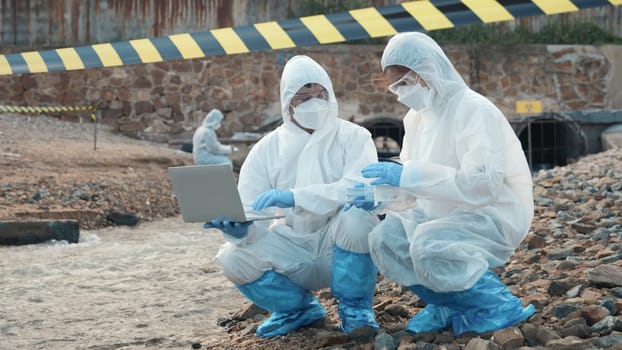 Ecologist sampling water from the river with test tube and save data to laptop computer, Biologist in a protective suit and mask collects sample of waste water from industrial, problem environment