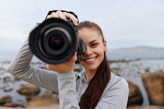 A cheerful young woman enjoys photography at the beach, holding a camera close to her face The soft colors of the ocean and rocks highlight her enthusiasm for capturing moments