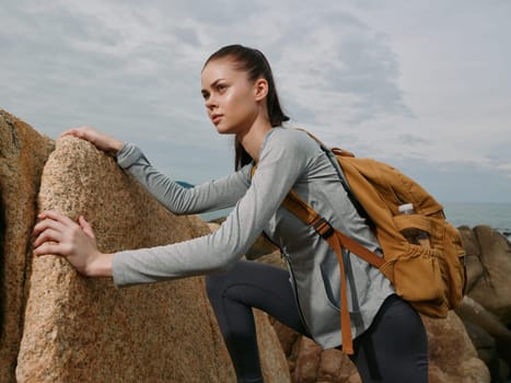 Determined young woman in gray activewear and a brown backpack climbs rocky terrain by the ocean, exemplifying adventure, endurance, and outdoor exploration