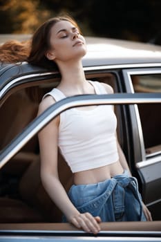 A cheerful young woman with a serene expression savors the warm sunlight outside a car door, dressed in a white crop top and denim, embodying summer freedom.
