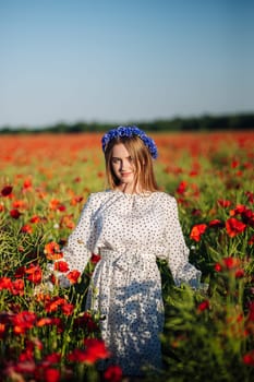 Beautiful woman wearing a blue cornflower wreath, posing gracefully in a vibrant red poppy field on a sunny summer day