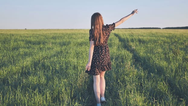 Young woman with long brown hair walks through a green field at sunset, her arm outstretched, enjoying the peace and beauty of nature