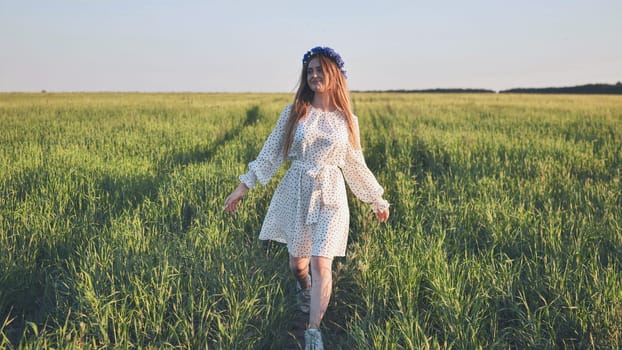 Beautiful young woman with long hair wearing a flower crown and white dress walks through a green field on a sunny summer day