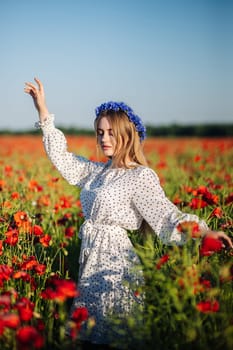 Beautiful blonde woman wearing a white polka dot dress and a blue flower crown, posing in a poppy field during a sunny summer day
