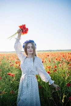 Woman wearing white dress and blue flowers crown holding poppy bouquet in a poppy field during a sunny summer day