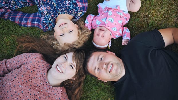 Cheerful parents with two children relaxing on green lawn, sharing laughter and connection during bright summer afternoon
