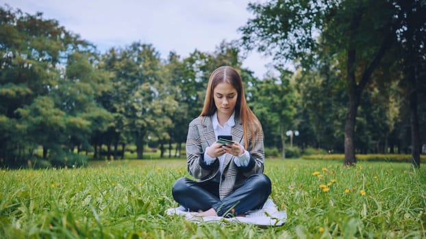 Professional working woman relaxing on grass, browsing mobile while holding credit card, blending technology and outdoor workspace seamlessly