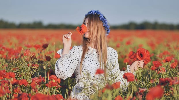 Blonde woman wearing floral crown inhaling fragrant red poppy amid blooming field during golden sunset light