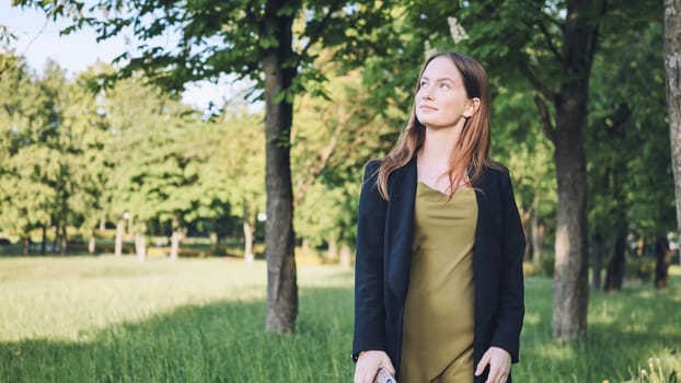 Professional woman strolling through verdant park during midday break, breathing deeply while gazing upward through leafy branches