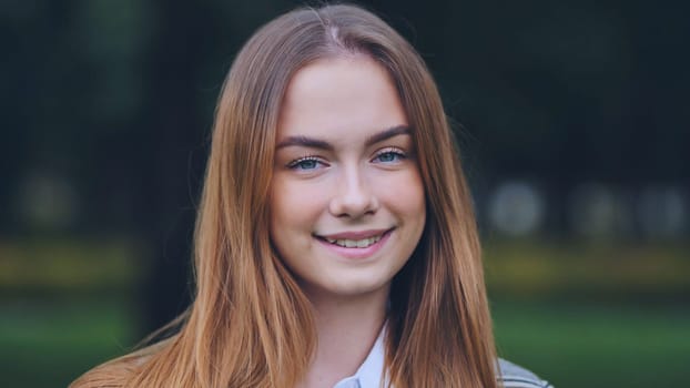 Teenage girl with flowing light brown hair, radiating joy while standing amid lush greenery of park landscape, expressing carefree youthfulness