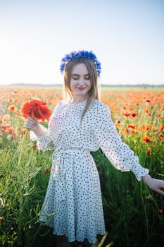 Smiling female wearing floral headpiece, white dotted sundress, carrying red poppy bouquet amid blooming meadow landscape