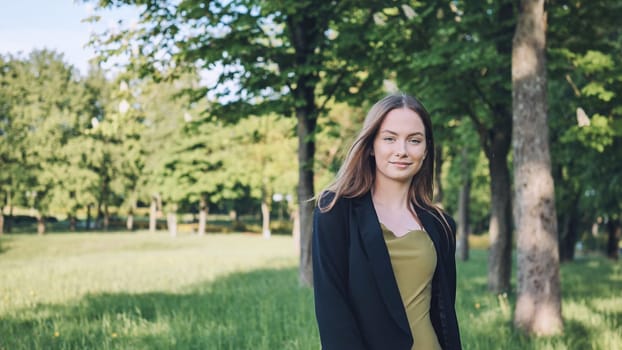 Professional businesswoman smiling, standing amid green urban park, basking in warm sunlight and natural surroundings