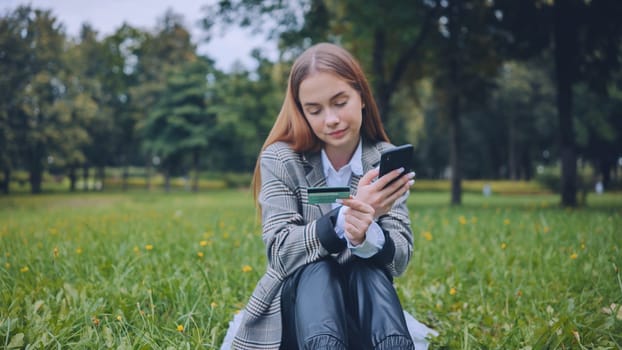 Young student paying online with credit card and mobile phone while sitting on grass in a park, enjoying convenient and secure mobile banking