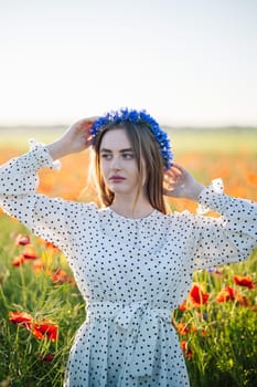 Slavic woman wearing cornflower wreath, white dress standing amid blooming poppy field during golden sunset light