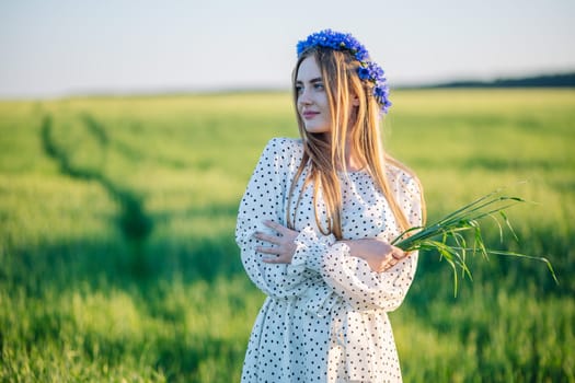 Blonde woman wearing cornflower wreath, holding wildflower bouquet, standing barefoot in lush green meadow, radiating summer freedom.