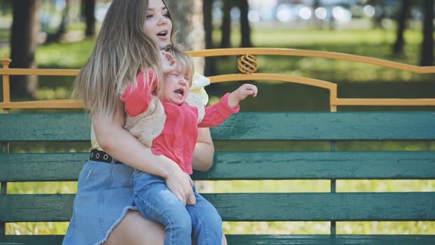 Young mother soothing her crying baby daughter while sitting on a bench in a public park, creating a moment of love and connection