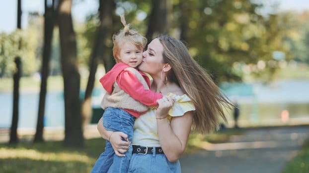 Young mother kissing her daughter on the cheek while joyfully holding her in her arms, surrounded by the beauty of a sunny park