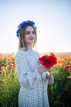 Beautiful woman wearing a cornflower wreath, holding a vibrant bouquet of red poppies while standing in a blooming poppy field