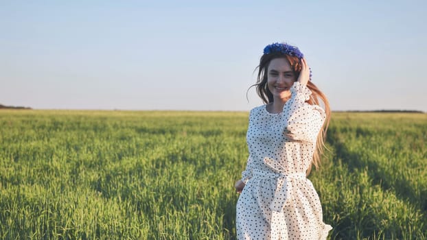 Beautiful smiling girl with blue cornflowers wreath running through green wheat field, enjoying freedom and nature during sunset