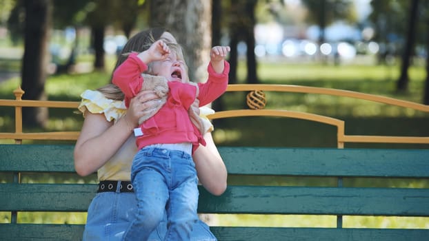 Mother holding and comforting her crying daughter while sitting on a bench in a park during a sunny day