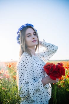 Beautiful blonde woman with cornflower wreath holding a bouquet of poppies in a field during a sunny summer day