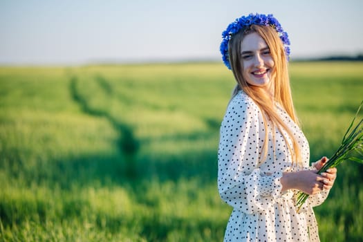 Young blonde woman with blue cornflowers crown holding bouquet of green ears in wheat field at sunset