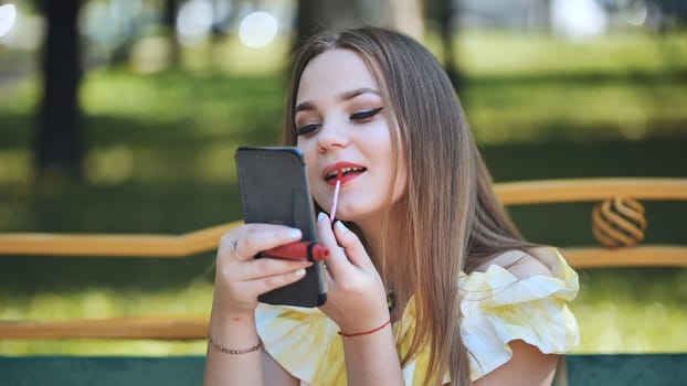 Young woman applying lipstick using her smartphone as a mirror while sitting on a bench in a park