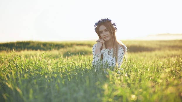 Beautiful young slavic woman wearing a flower wreath sitting in a green field at sunset, enjoying the peaceful atmosphere