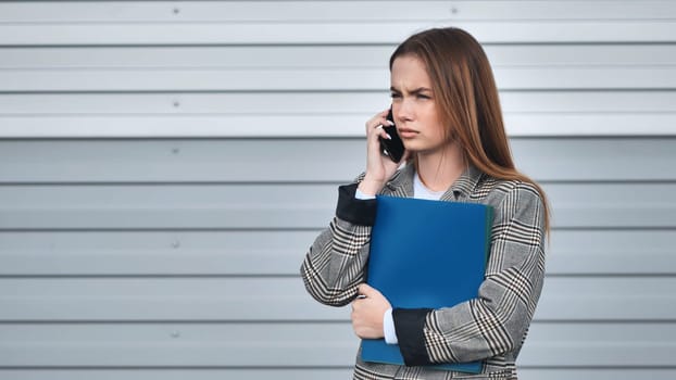 Young businesswoman holding a folder and making a phone call with an annoyed expression, standing in front of a metal wall